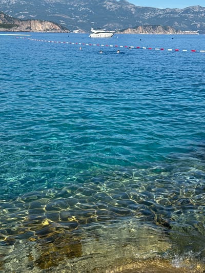 The image shows a clear blue sea with visible rocks and sand beneath the surface. The water is calm with gentle ripples. In the distance, a white yacht is anchored, and a few people are swimming in the sea. A rope with red and white buoys separates a swimming area from the open water. Beyond the sea, a mountainous coastline is visible, with some buildings scattered on the slopes. The sky is clear and blue. The location is Prijevor, Montenegro.