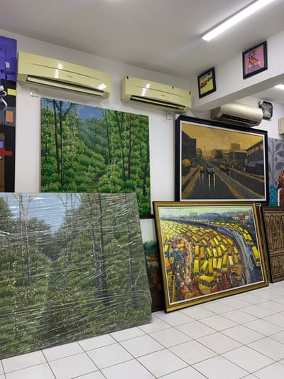 An interior view of an art gallery or storage space in Ikate, Nigeria, displays multiple artworks. The room features white walls and a white tiled floor with a grid pattern. Three cream-colored wall-mounted air conditioning units are visible, two with "Panasonic" branding. White fluorescent strip lighting illuminates the ceiling. On the left wall, a large unframed painting depicts a dense green forest. Below it, a large landscape painting, similar in composition, is wrapped in clear protective plastic and leans against the wall. A prominent framed oil painting, depicting a city street scene with buildings, vehicles, and wet asphalt under a muted sky, hangs centrally on the right wall. Below this, another large framed painting, rendered in an Impressionistic style, depicts an elevated perspective of a winding road congested with numerous yellow vehicles, resting on the floor. To its right, a dark, intricately carved wooden panel leans against the wall. Higher on the walls, several smaller framed artworks are visible, including one with abstract figures and another with a yellow background. No individuals are present in the scene.