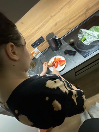 A person is seen from a high angle view, chopping red bell peppers on a white plate with a knife. They are wearing a black and beige patterned shirt. The scene appears to be in a modern kitchen with a wooden countertop, a black induction stovetop, and various kitchen appliances, including an electric kettle. A smartphone and a plastic bag with a green vegetable, possibly a cucumber, are also visible on the countertop. The location is identified as Kommunarka, Russia.