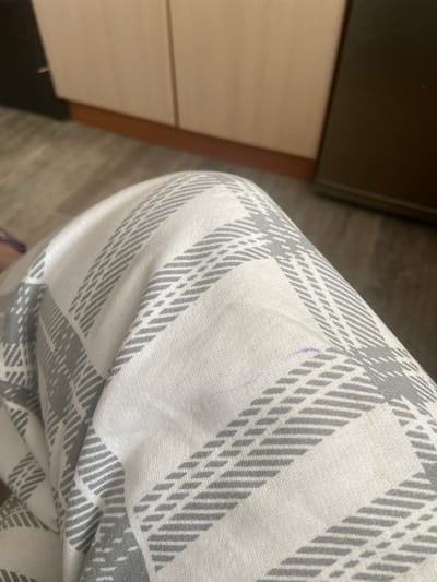 The media file shows a close-up view of a person's leg wearing plaid pajama pants. The pants are white with gray plaid markings. A faint purple mark, possibly from a pen, is visible on the fabric. In the background, the edge of a dark brown furniture piece and a light-colored drawer unit are discernible, suggesting an indoor setting, likely a bedroom. The floor appears to be made of wood or laminate flooring. The lighting is neutral, indicating it could be daytime or the room is well-lit. There is no text visible in the image. The context provided places the location as Karpinsk, Russia.