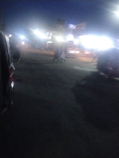 The image depicts a highly blurry and dark night scene on a street, likely in an urban or semi-urban environment. The primary subjects are numerous bright, overexposed artificial light sources, presumably from vehicle headlights, streetlights, and possibly illuminated buildings or signs. These intense lights create significant glare and lens flare, obscuring much of the detail.
In the immediate foreground on the left, a portion of a vehicle, displaying red and dark colors, is visible, suggesting the perspective is from within or very close to another vehicle. The ground beneath is a dark, indistinct surface, likely a road or paved area.
Faint, silhouetted forms of several people can be vaguely discerned in the middle ground, appearing to walk or stand amidst the vehicles. Their features and specific activities are entirely obscured by the blurriness and low light conditions. The presence of multiple light sources and indistinct forms suggests general traffic and movement on a busy street. No legible text is visible due to the image quality and overexposure, though some faint markings might exist on distant, illuminated structures. The overall impression is that of a bustling street scene in Uyo, Nigeria, after dark, characterized by a lack of clarity and an abundance of bright, diffused light.