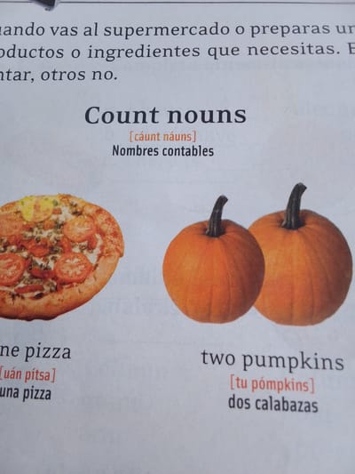 The image displays a page from what appears to be a language learning book or material. The main subjects are food items and a grammatical concept. On the left, a whole pizza is pictured, and below it, the text "one pizza [uán pítsa] una pizza" is visible, indicating this is an example of a "count noun" in English and its translation into Spanish. On the right, two pumpkins are shown, accompanied by the text "two pumpkins [tu pómpkins] dos calabazas," further illustrating the concept of count nouns.

Above these examples, the heading "Count nouns" is prominently displayed in large, bold font, with the phonetic pronunciation "[cáunt náuns]" and the Spanish translation "Nombres contables" below it.

In the background, there is text in Spanish that seems to be part of a lesson or instructional material. This text reads: "ando vas al supermercado o preparas ur odúctos o ingredientes que necesitas. E ntar, otros no." This translates to something like: "When you go to the supermarket or prepare something, you need products or ingredients. You take some, others not."

The setting appears to be an indoor environment, likely a classroom or study space, given the presentation of educational content. There are no people visible in the image. The lighting is even, suggesting an indoor setting during the daytime. The overall mood is educational and informative. The location context of La Tagua, Venezuela, is not directly indicated by any visual cues within the image itself.