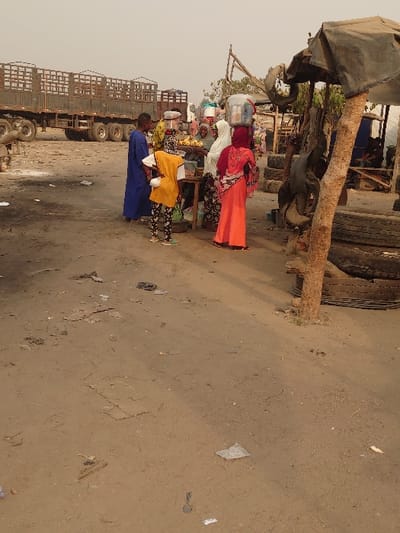 The image depicts an unpaved outdoor area in Ajaokuta, Nigeria, characterized by dry, light-brown earth and scattered debris including paper and plastic. In the midground, a group of approximately six individuals is gathered around a low wooden vendor stall displaying yellow and green produce. One person behind the stall, wearing a white head covering, attends to the goods. Among those facing the stall are an individual in a long blue garment, a young person in a yellow top and patterned leggings, and another individual in a reddish-orange patterned dress with a matching head covering, all seen from behind. Two individuals further back are observed carrying containers balanced on their heads. In the background to the left, two dark-colored, multi-axle flatbed trucks with metal railing structures are parked. To the right, a makeshift shelter constructed from a tree trunk and a dark fabric tarp stands near a stack of used vehicle tires.