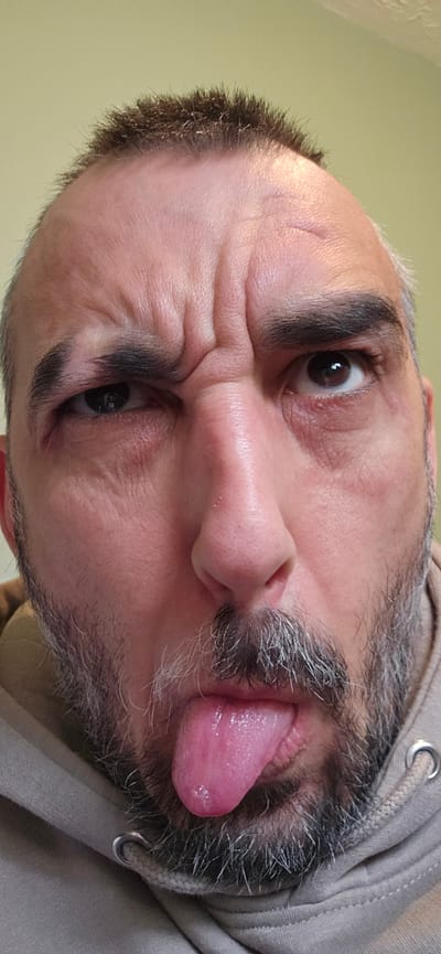 A close-up, rotated portrait captures an adult male in Norwich, United Kingdom, making a playful or silly expression. His head is tilted, and he is looking upwards and to the right within the frame. He has dark, short hair and a short beard or stubble, both showing noticeable streaks of gray. He is sticking his pink tongue out to the left side of his face and scrunching his eyes and forehead, creating pronounced wrinkles across his brow and around his dark eyes. He is wearing a light-colored hooded sweatshirt, with the fabric and a metal drawstring grommet visible on the left side. The lighting appears soft and natural, suggesting it is daytime. The background is a plain, out-of-focus, light-colored wall, providing no specific location cues other than an indoor or simple setting. No text is visible in the image.
