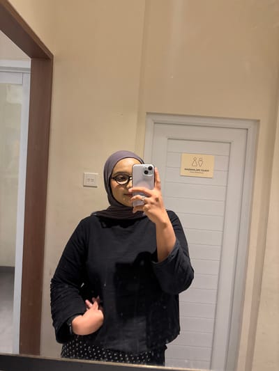 A young woman wearing a grey hijab and black long-sleeved shirt is taking a mirror selfie. She is also wearing glasses and a polka dot skirt. The background shows a wall with a door and a sign indicating it is a toilet. The image appears to have been taken in a restroom.