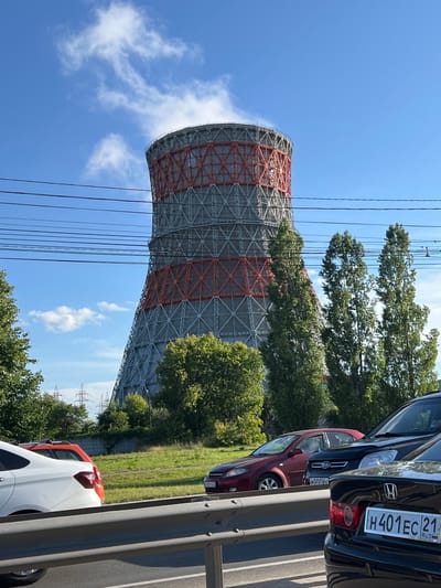 A wide shot captures a cooling tower, a large industrial structure, against a clear blue sky dotted with wispy clouds. The tower is cylindrical, with a lattice-like exoskeleton painted in red and gray sections. Some of the upper portion of the tower is obscured by trees, while a plume of white vapor emerges from its opening, suggesting it is in operation.
In the foreground, three cars are parked. The closest is a white car, partially visible, followed by an orange car, and then a dark red car. Further down the line is a black car, the rear of which is clearly visible. The license plate of the black car reads "H 401EC 21 RUS". The cars are parked alongside a metal barrier, indicating a roadside or parking area.
The scene is set amidst lush green trees and vegetation. In the background, power lines and electrical pylons are visible, hinting at the industrial or urban nature of the location. The lighting suggests it is daytime, likely late morning or afternoon, with bright sunlight casting shadows. The overall atmosphere is one of a functional industrial environment integrated with nature. The image is oriented vertically, with the cooling tower dominating the left side of the frame and the cars and roadside on the right. The perspective is looking upwards towards the top of the cooling tower.