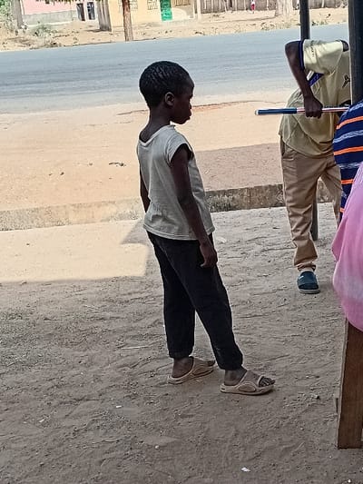 The image depicts a daytime scene in Dabawa, Nigeria, featuring a young child and partially visible adults on a dirt ground adjacent to a paved road. In the foreground left, a child with dark skin and short, dark hair wears a light-colored sleeveless t-shirt, dark long pants, and light brown open-toed sandals. The child is positioned facing slightly right, with head turned further right, observing something outside the frame. To the child's right, an adult male, partially visible and bent over, wears a light-colored short-sleeved collared shirt, light brown pants, and dark blue closed-toe shoes. This adult holds a long, slender object with blue, white, and red sections. Another individual, partially visible to the right, wears a blue and orange striped shirt. Further right, a section of pink fabric and a wooden structure are present. The immediate ground is dirt, transitioning to a concrete curb, and then a gray asphalt road. Across the road, multiple single-story buildings, some light-colored and one with a prominent green door, are visible under bright lighting conditions.