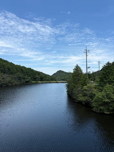The image shows a serene landscape with a body of water in the foreground, flanked by lush green hills and trees. The water is dark blue with subtle ripples, reflecting the blue sky above, which is dotted with scattered white clouds. On the right side of the water, a line of trees and vegetation meets the water's edge, and beyond that, there are utility poles with electrical wires stretching across the scene. The background features rolling hills covered in dense green foliage, with a faint misty atmosphere in the distance. The overall impression is one of natural beauty and tranquility, possibly a reservoir or a large pond within a hilly, forested area. This scene is located in the city of Naoshima, Japan, a place known for its art islands and natural landscapes.