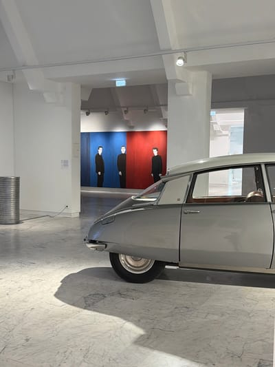 The image captures an indoor exhibition space in Marseille, France, likely an art gallery or museum. In the right foreground, a light grey classic Citroën DS sedan is stationary, positioned diagonally with its rear passenger side prominently displayed. The vehicle features a lighter, possibly white, two-tone roof and visible tan interior upholstery.

To the left, a tall, cylindrical sculpture composed of stacked, corrugated metallic discs stands on the polished light grey marble floor, which exhibits distinct dark veining.

Through a central architectural opening, an art installation is visible in the background. This installation comprises three large, vertical rectangular panels: two blue panels on the left and one red panel on the right. Each panel features a full-body, minimalist depiction of a man dressed in black, facing forward.

The environment is characterized by stark white walls, columns, and ceilings, illuminated by track lighting. Shadows from the car and architectural structures are cast across the reflective marble floor. In the far background, beyond the art installation, a brightly lit hallway or room containing stairs is faintly visible through another opening. No living individuals are present in the scene.