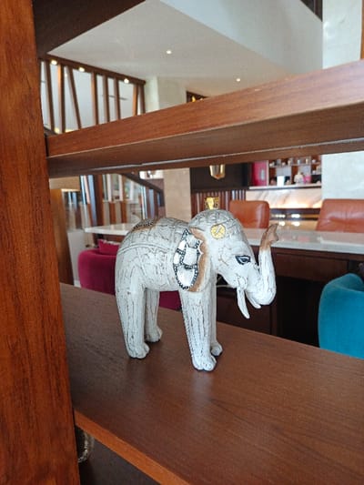 An ornate white elephant figurine sits on a wooden shelf. The shelf is part of a larger wooden structure that dominates the foreground. In the background, a bright interior is visible, featuring light-colored walls, patterned dividers, and furniture in various colors. Some chairs are upholstered in pink and orange, with a blue chair nearby. The scene is well-lit, creating a warm and inviting atmosphere.
