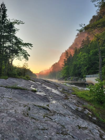 The image depicts a tranquil natural scene, likely at sunrise, with a body of water flowing towards a dam or spillway. The foreground is dominated by a large, grey, textured rock formation with patches of green moss and grass. To the left, a dense cluster of tall green trees rises against a pale, hazy sky. The background features a steep, tree-covered slope, bathed in the warm, orange glow of the rising sun, with a mist or fog adding a soft diffusion to the light. A yellow buoy is visible in the water. On the right side of the image, a concrete structure with a white railing, presumably part of the dam or a viewing platform, is situated at the edge of the water. The overall impression is one of serenity and the beauty of untouched nature. The prompt references a specific location in Saint-Alban, Canada, but this information cannot be visually verified from the image alone.