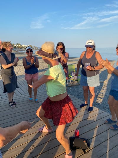 The image depicts a group of people on a wooden boardwalk next to a beach. The sun is low in the sky, casting long shadows, indicating it is either early morning or late afternoon. The weather appears to be clear and sunny, with a bright blue sky and a few wispy clouds.
There are at least six individuals visible, appearing to be in their 40s or older. Most are casually dressed, with some wearing hats and sunglasses, suggesting a relaxed, possibly vacation or social gathering. One person wears a white baseball cap with "Los Angeles" visible on it. Another person is wearing a wide-brimmed straw hat. The clothing styles include shorts, tank tops, and dresses, suitable for warm weather.
The group seems to be interacting, with some individuals clapping or gesturing. One person is in a dynamic pose, appearing to be reaching out or stepping. Another person is holding a clear plastic cup. In the background, the sandy beach and the ocean are visible, with some palm trees and buildings suggesting a coastal resort town, consistent with the Marbella, Spain location context. There is also a large black portable speaker and a red cylindrical object on the boardwalk. The overall mood appears to be cheerful and convivial.
