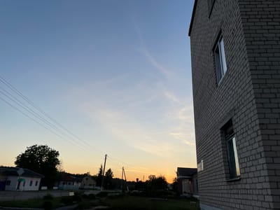The image depicts a rural scene at dusk in Азершчынскі сельскі Савет, Belarus. A tall, gray brick building with windows occupies the right side of the frame, casting a shadow. In the distance, a row of houses and trees are visible against a sky transitioning from blue to a warm orange glow near the horizon. Power lines stretch across the sky, supported by utility poles. The ground in the foreground is covered in grass and vegetation. There are no people or animals clearly visible in the image.