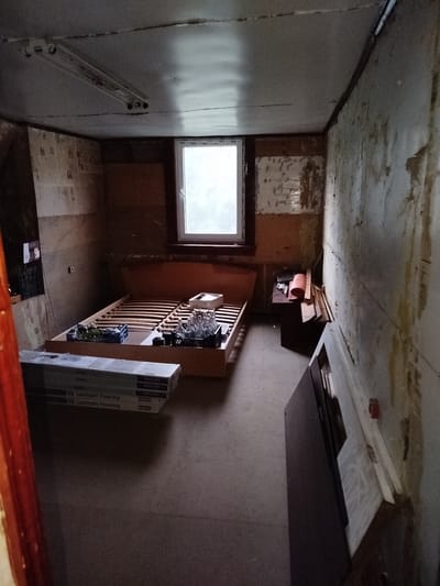 The image depicts an unfinished room with a bed frame and various items scattered around. The walls are unfinished, showing patches of peeling wallpaper and exposed plaster. A window is visible on the back wall, letting in natural light. On the bed frame, there are plastic crates containing bottles and other objects. To the left of the bed, stacks of laminate flooring are leaning against the wall. A small wooden desk with items on it, including a rolled-up mat and some wooden planks, is positioned near the window. In the foreground, part of a wooden doorway is visible. The floor is concrete. The scene appears to be in a state of renovation or disarray. The image was taken in Дзешчанскі сельскі Савет, Belarus.