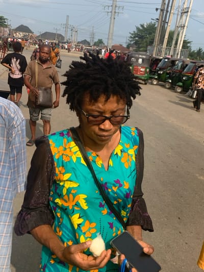 This image depicts an outdoor urban scene in Ura Nta Umuarandu, Nigeria, during daylight hours. In the foreground, a female subject with dark dreadlocks and dark-rimmed glasses wears a teal top adorned with orange, yellow, and purple floral patterns, featuring dark, long sleeves. She holds a small, white, plastic-wrapped spherical object in her left hand and a black smartphone in her right, with her head oriented downwards. To her immediate left, a male is partially visible, wearing a light blue and white checkered shirt.
In the midground, a male with short dark hair, dressed in a brown polo shirt, grey shorts, and dark flip-flops, carries a dark leather messenger bag across his left shoulder and looks forward. Several other pedestrians are scattered throughout the midground and background, indicating a populated area. The background features an asphalt road where a line of green and red auto-rickshaws, identified as "Keke Napep," are parked on the right side. Overhead, numerous utility poles and an intricate network of electrical wires are visible against a bright sky. Distant buildings with light-colored roofs complete the urban landscape.