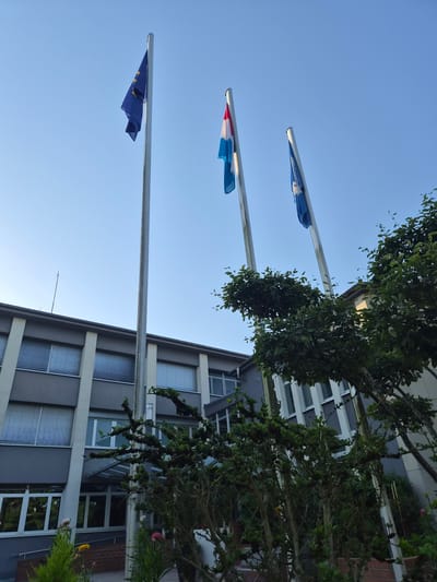 The image depicts the exterior of a building, identified as the Sanem town hall in Luxembourg. The building has a modern architectural style with a grey facade, white vertical accents, and large windows covered by horizontal blinds. In front of the building, three flagpoles are visible, each flying a flag. The leftmost flag is the European Union flag. The middle flag appears to be the flag of Luxembourg. The rightmost flag is blue with white text and symbols, possibly representing an organization or a regional flag. Dense green foliage, including trees and bushes, partially obscures the lower part of the building and the flagpoles. The sky is clear and blue, suggesting it is daytime. The scene is static and shows no people.