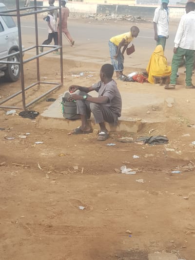 The image shows a street scene in what appears to be Kufena, Nigeria. A man is crouching near a backpack, possibly working on something with small objects. Several other people are walking along the road in the background. The surrounding area is a mix of paved and unpaved ground, with litter scattered about. A metal frame structure is in the foreground, partially obscuring a vehicle.