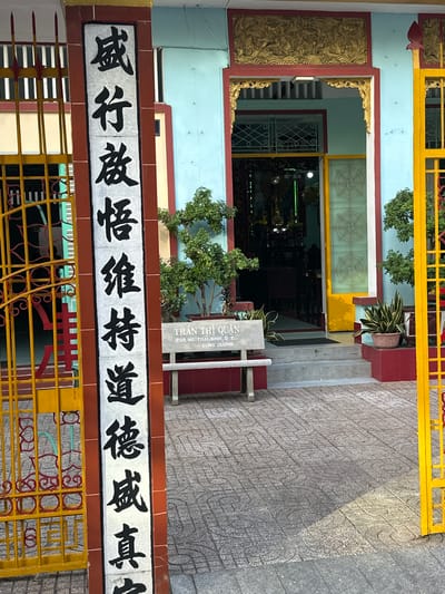 The image shows a brightly colored building, likely a temple or a place of worship, in Thủ Đức, Vietnam. The building features a light blue facade with red and yellow accents. A yellow metal gate with intricate red decorative elements is in the foreground. To the left, a gilded relief panel adorns the wall, depicting intricate patterns.
In the center of the image, an entrance to the building is visible, framed by red and gold trim. Through the entrance, the interior appears dimly lit, with glimpses of statues and possibly religious artifacts. A stone plaque near the entrance displays Vietnamese text, reading "TRẦN THỊ QUẬN 209.NG. THAI. ΒΙΝΗ.Ο.Σ CÙNG DƯƠNG". Above the entrance, a horizontal banner displays large black Chinese characters on a white background, bordered by red. The characters are stylized and appear to be a religious or philosophical phrase.
Lush green trees are planted in pots near the entrance, and more trees are visible around the building, suggesting a well-maintained outdoor space. The ground in front of the building is paved with stone tiles featuring a repeating pattern. The lighting suggests it is daytime, possibly late morning or afternoon, casting shadows on the paving stones. The overall scene conveys a sense of cultural and religious significance.