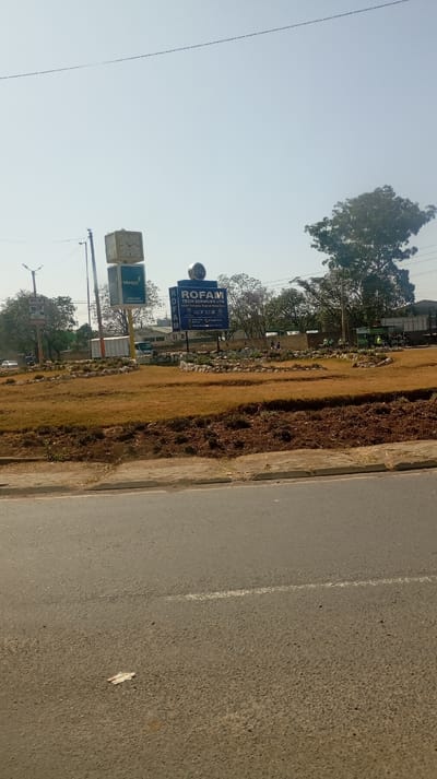 An outdoor scene features a street, median, and sky. In the median, brown vegetation and freshly tilled dark soil surround a round garden area with a sign for "ROFAM TECH SERVICES LTD." A billboard-like clock with the Stanbic Bank logo is visible to the left. Trees are scattered in the background, and utility poles with power lines extend across the top of the frame. A single leaf lies on the asphalt of the street.