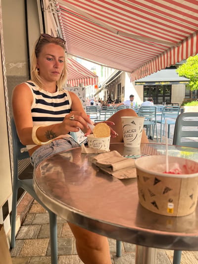 A young woman with blonde hair is sitting at a table outdoors, possibly at a cafe. She is wearing a striped top and denim shorts. She is holding a cone with an ice cream cone on top, and there is another ice cream cup on the table. The scene is set under a red and white striped awning, and in the background, there are tables and chairs, and some buildings with awnings. The sunlight suggests it is daytime, and the overall atmosphere appears relaxed and casual. There is text visible on the ice cream cup: "GLACIER VILFEU" and "CANNES".