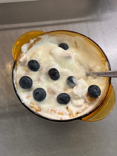 A close-up, overhead shot shows a golden-orange glass bowl filled with yogurt and blueberries. Several whole blueberries are scattered across the creamy white yogurt. A metal spoon is partially submerged in the yogurt on the right side of the bowl. Small pieces of what appears to be granola or cereal are visible within the yogurt. The bowl sits on a textured grey surface, possibly a countertop. The background is out of focus, showing horizontal lines that could be shelving or a wall. The location is not specified in the image or metadata.