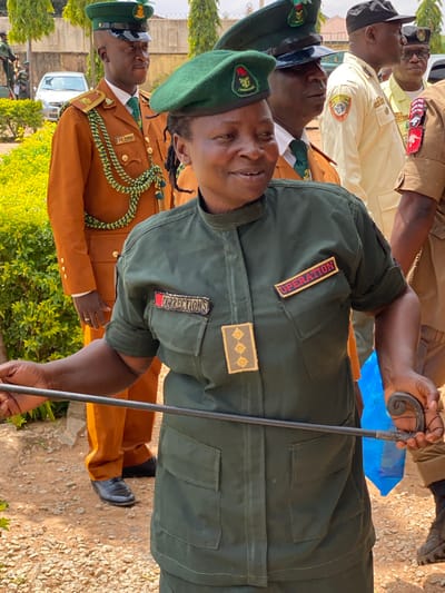 The image shows a group of people, likely correctional officers, in uniform. The primary subject is a man in the foreground wearing a dark green uniform with a green beret. He is holding a black baton-like object with a decorative metal handle. Patches on his uniform read "OPERATION" and "CORRECTIONS". He appears to be looking slightly up and to the left, with a neutral expression.
Behind him, other individuals are visible in various uniforms. To his left, a man in a lighter brown uniform with a green and yellow cord across his chest looks towards the camera with a slight smile. Another man, further back and to the left, wears a light yellow uniform and looks upwards. To the right of the primary subject, a person in a brown uniform is partially visible, also holding a similar baton. The setting appears to be outdoors, with gravel on the ground and some greenery in the background. The lighting suggests it is daytime, possibly morning or afternoon, with mild weather. The presence of uniforms and official-looking attire points to a formal event or duty. The image is oriented vertically.