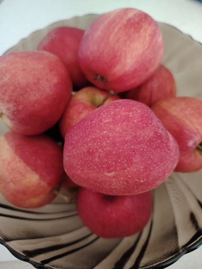 A close-up, overhead shot features a shallow, dark glass bowl filled with a pile of bright red apples. The apples are a vibrant red, with some showing hints of yellow and faint streaks of lighter red. The bowl's textured, ribbed surface is visible at the bottom and right side of the image, creating a subtle play of light and shadow. The background is a plain, light-colored surface. There are no people, actions, or specific environmental details that would suggest a location in Кривец, Russia.