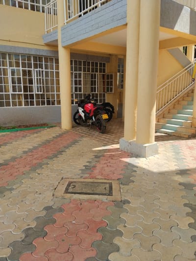 The scene depicts an outdoor paved area adjacent to a multi-story, light-yellow building, located in Hospital ward, Kenya. The building's ground floor features multiple large windows with white frames and numerous small panes. An upper floor is visible with a balcony guarded by white railings and a horizontal grey decorative band below it. On the right, an exterior staircase with white railings ascends. Yellow-painted cylindrical columns support an overhang. The ground is paved with interlocking concrete pavers in alternating patterns of reddish-brown, light beige, and dark grey. A rectangular metal utility access panel is embedded within the paving. Two motorcycles are parked beneath the building's overhang. The more prominent one is a red and black street bike, displaying a Kenyan license plate "KMER 932Y". A second, darker motorcycle is partially visible behind it. Strong sunlight illuminates the scene, casting distinct shadows on the paved ground and building facade. A narrow strip of green vegetation is visible at the base of the building wall on the left. No individuals are present.