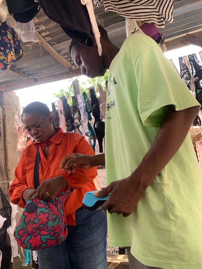 Two people, likely a mother and child, are looking at a display of bras and other clothing items at an outdoor market. The bras are hanging from a makeshift rack made of corrugated metal and wood. The background suggests a sunny day in a tropical or subtropical climate, with trees visible in the distance. The people appear engaged with the merchandise, possibly considering a purchase. The scene is reminiscent of a typical market setting in Nyanya, Nigeria, where such stalls are common. There is some visible text on a sign in the background, but it is mostly illegible due to its small size and resolution. The overall impression is a snapshot of everyday life at a bustling local market.