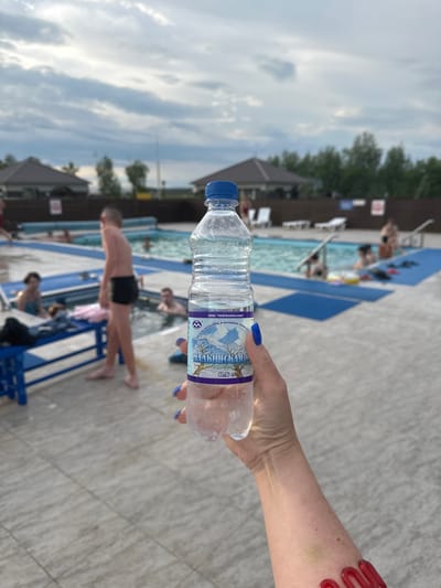 In Yuzhnyye Koryaki, Russia, a person holds a bottle of mineral water in the foreground, with a swimming pool and surrounding amenities visible in the background. The pool area features blue mats and lounge chairs, and several people are in and around the water. The sky above is overcast. The image captures a leisure scene at an outdoor pool.