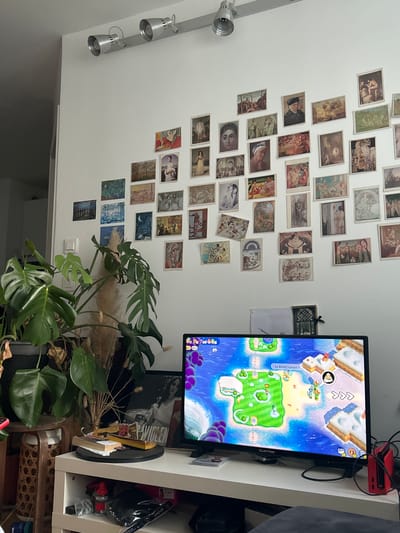 The photograph depicts an interior scene in Pantin, France, featuring a wall adorned with numerous small prints or postcards, arranged in a somewhat haphazard manner. To the right, a television displays a colorful video game interface, suggesting a playful or entertainment-oriented atmosphere. A large Monstera plant occupies the lower center of the frame, adding a touch of nature to the indoor environment. The television is positioned on a white shelving unit, which also contains various items, including books and other objects, contributing to a sense of lived-in clutter. Overhead track lighting illuminates the scene.