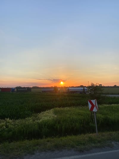 This media file captures a sunset scene in Podłęże, Poland, featuring several large trucks parked along a grassy roadside next to a paved surface. The sun is low on the horizon, casting a warm, golden light across the sky, which transitions from a soft blue to hues of orange and pink. Wispy clouds are visible, catching the light of the setting sun.
Among the trucks, one is a white tanker truck with text that reads "www.pol-bauschomer.com". Another truck is a dark green box truck with the text "DUVENBECK" printed in white letters. A red container is also visible behind one of the trucks. A prominent feature is a roadside sign with red and white diagonal stripes, indicating a warning or a hazard.
The setting appears to be rural, with lush green grass and vegetation bordering the road and the area where the trucks are parked. The sky indicates it is late afternoon or early evening. The overall mood conveyed by the image is one of tranquility, with the beauty of the natural sunset contrasted by the industrial presence of the trucks.