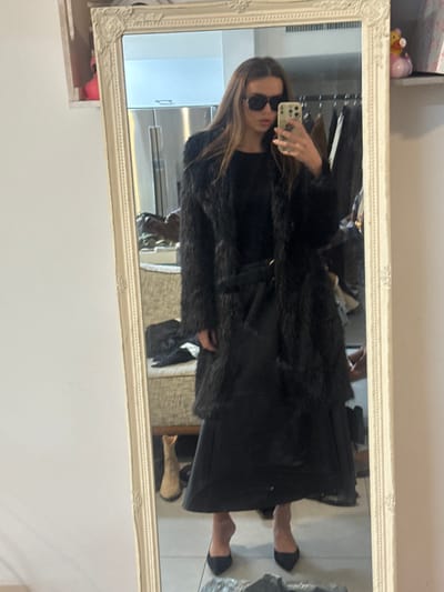 This image depicts a full-length mirror selfie of a female individual, taken within an interior space in the city of Kfar Yona, Israel.
The subject, a female with long, straight brown hair, stands facing an ornate, off-white framed mirror. She wears dark, rectangular sunglasses and is dressed in a black ensemble consisting of a long, shaggy or faux fur coat, a dark long-sleeved top, a flowing black midi-skirt, and black pointed-toe high heels. Her right hand, partially obscured by the sunglasses, holds a light-colored smartphone, positioned to capture her reflection.
The background, visible in the mirror's reflection, reveals a domestic setting. To the left, a white wall is visible, with a small shelf near the top displaying a pink figurine. To the right, a chrome-finished appliance, possibly a refrigerator, is present. Further right, a clothing rack with various dark garments hangs, and an upholstered, light-colored chair or sofa is partially visible. On the floor in the background, a light brown boot is discernible. The overall environment suggests a room within a residence, likely a bedroom or dressing area, characterized by personal items and clothing.