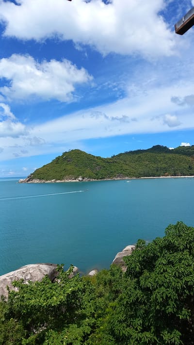 The image depicts a tropical coastal scene, oriented vertically. The foreground on the left features a section of a metallic structure, possibly part of a building or observation deck, against a bright blue sky filled with scattered white clouds. The majority of the image is dominated by a body of clear, turquoise water. Along the waterline, a lush, green, tree-covered island or peninsula juts out into the water. The island is characterized by a steep, mountainous terrain blanketed in dense vegetation. A narrow strip of beach is visible along the edge of the island. Further out in the water, a small boat is leaving a white wake, indicating movement. The time of day appears to be daytime, with clear and sunny weather suggested by the bright sky and the intensity of the light. The scene evokes a sense of natural beauty and tranquility. The location is identified as Ban Tai Subdistrict, Thailand, which aligns with the tropical island imagery. No visible text is present in the image.