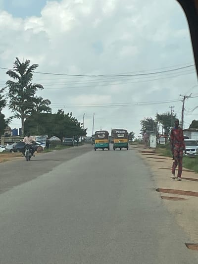 The image shows a street scene in Gwong, Nigeria. The foreground features a paved road with visible potholes. On the right side of the road, a man in a red and black patterned garment is walking away from the camera. Further down the road, two yellow and green auto-rickshaws are parked. On the left side of the road, a man is riding a motorcycle, approaching the viewer. Several vehicles are parked or moving along the road, and trees line the sides of the street. The sky is overcast with clouds. The overall impression is of a typical day in a Nigerian town.