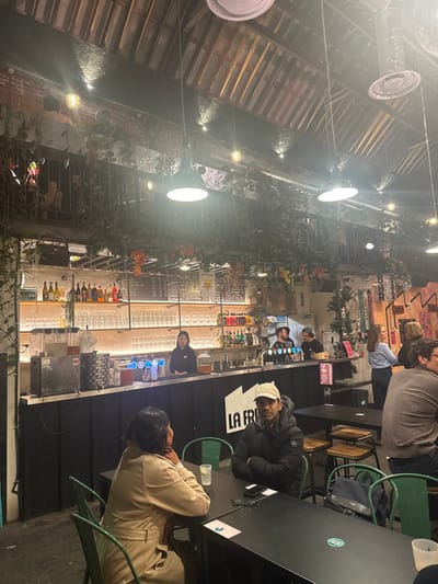 This image depicts the interior of a commercial establishment, likely a bar or restaurant, located in Saint-André-lez-Lille, France. The space features a high, pitched ceiling constructed with exposed wooden beams, illuminated by numerous industrial-style pendant lights and spotlights. Artificial green foliage and vines are extensively draped across the ceiling structures, a mezzanine railing, and above the bar area, creating a verdant effect against an industrial backdrop.
In the foreground, two individuals are seated at a black square table with green metal chairs. A woman, with dark hair and wearing a light beige trench coat, is positioned with her back partially to the camera, her right arm resting on the table beside a glass containing a lime wedge. Opposite her, a man with dark skin, wearing a black puffer jacket and a light-colored baseball cap, is seated facing the camera with his arms crossed, looking directly into the lens. A smartphone and sunglasses are placed on the table in front of him.
The main bar counter, a long, black structure with corrugated detailing on its front, stretches across the mid-ground. Partial white lettering, "LA FRI", is visible on the bar's facade. Behind the counter, an East Asian female bartender wearing a black hoodie stands attentive. To her right, two male staff members, one bearded, are visible operating beer taps and preparing drinks. Illuminated shelves behind the bar display various liquor bottles and multiple rows of clean glassware. Menu boards with text and graphics are mounted above these shelves.
Throughout the mid-ground and background, additional patrons are visible, seated at other black tables or standing near the right end of the bar, socializing. A small artificial Christmas tree is positioned near the bar's right extremity. On a mezzanine level above the main floor, several indistinct figures are present behind a railing also adorned with greenery. The visible walls consist of exposed brickwork. The entire scene is brightly illuminated by artificial lighting, indicating an indoor evening or night setting.