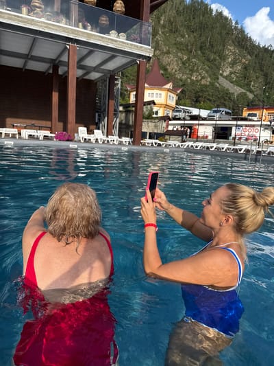 In Barangol, Russia, two women are in a swimming pool. The woman in the foreground on the right, facing left, has blonde hair pulled into a bun and is wearing a blue and white swimsuit. She is holding a pink smartphone and appears to be taking a photo or video. She is wearing a red bracelet on her left wrist and a pearl necklace. The woman in the foreground on the left is facing away from the camera, with blonde hair, and is wearing a red swimsuit. She is partially submerged in the water. The pool water is clear and blue. In the background, there is a building with wooden pillars and a balcony adorned with potted plants. Further back, there are lounge chairs, a yellow building with a red roof, and a forested mountainside under a blue sky with clouds. A sign with Russian text and a logo is visible in the background.