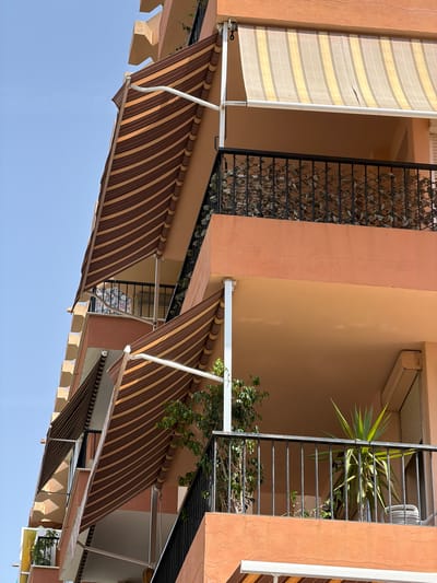 The image is an outdoor, low-angle shot of an apartment building with multiple balconies and retractable awnings in Palma de Mallorca, Spain. The building's facade is painted a warm, peachy-orange color. Several striped awnings are visible, some extended and others retracted. One extended awning features brown and tan stripes, while another higher up has lighter tan and white stripes. The balconies have black metal railings, and one balcony on the right contains a large yucca plant and a smaller tree-like plant. Another balcony further down on the left has a patterned cloth hanging on its railing. The sky is a clear, bright blue. The architectural style suggests a Mediterranean setting.