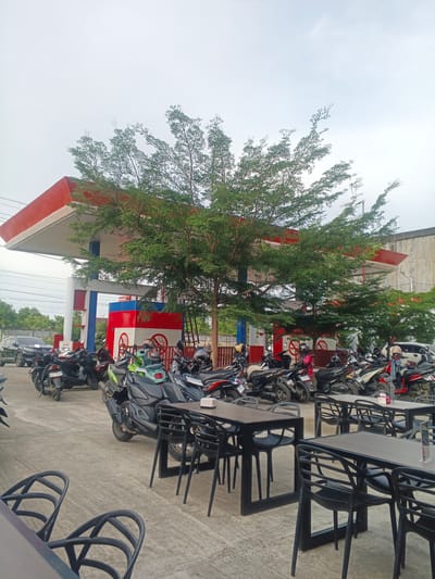 This image captures an outdoor scene at a gas station, likely in Geulumpangsulu Timu, Indonesia. The main subjects are numerous motorcycles parked in rows, suggesting a busy day or a popular gathering spot. A red and white gas station canopy dominates the background, with its distinctive pillars and roofing. Several trees with dense green foliage are strategically placed, providing shade over the parking area and the outdoor seating.
In the foreground, several black tables and chairs are set up, indicating an area for customers to rest or dine. The tables have small items on them, possibly napkins or condiments. The ground is paved with concrete.
The weather appears overcast, with a bright, diffused light suggesting it could be morning or late afternoon. The overall mood is functional and communal, with the gas station and seating area serving as a hub for locals and travelers.
Visible text includes a "PERTAMAX" sign on the gas station structure, which is a brand of fuel in Indonesia. There are also signs with red circles and diagonal lines, likely indicating prohibitions, such as "No Smoking." The license plates on some of the motorcycles are partially visible, showing alphanumeric characters typical of Indonesian plates. There are no prominent people visible, but the presence of parked motorcycles and seating implies human activity.