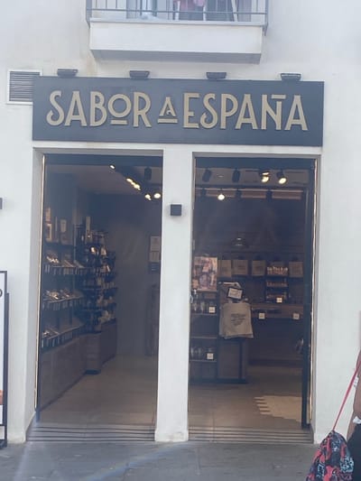 A sign above a storefront reads "SABOR A ESPANA" in gold lettering. The storefront features two entrances, one on the left and one on the right, revealing shelves stocked with various items, possibly food products or souvenirs. The left entrance shows a well-lit interior with displays of goods. The right entrance is also lit, showing more shelves and a counter area. The building appears to be made of white stucco. The location is Conil De La Frontera, Spain.