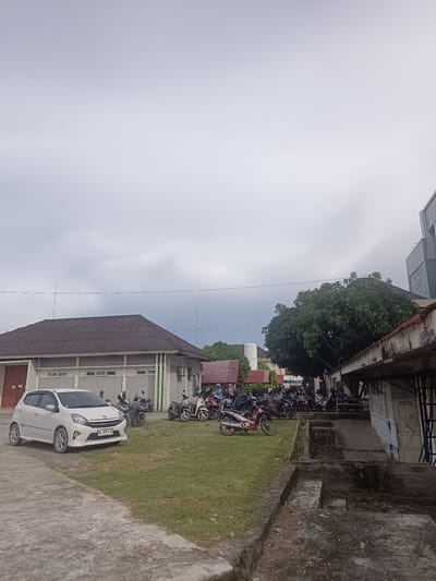 This is a medium shot, outdoor, eye-level, vertical photograph taken on a cloudy day. The scene is a parking area with several buildings and many parked motorcycles and one white car. In the foreground, a concrete path leads to a patch of green grass.

The main subjects are the parked vehicles and the buildings. A white hatchback car is parked on the left, facing the viewer. Behind it, a row of motorcycles is parked on the grass, some with riders sitting on them, suggesting a brief stop or wait. Further back, other buildings are visible, including a long, low-rise structure with a dark, corrugated roof and a modern building with a light-colored facade. A large, leafy tree stands prominently in the middle ground. The sky is overcast, casting a diffused light over the scene.

The activity appears to be casual, with people parked and gathered around their motorcycles. The time of day is likely daytime, given the ambient light, but the cloudy conditions might obscure the exact time. The presence of many motorcycles and the parked car suggests this is a public area or a place of gathering. No specific emotional cues are evident.

Visible text includes the license plate of the white car, which reads "BL 1892 AI". Another sign on a building in the background appears to have the word "SAM" as part of its text. A sign on a building in the far right appears to say "RUANG CUCI". The location context indicates Lampeudeu Baroh, Indonesia.