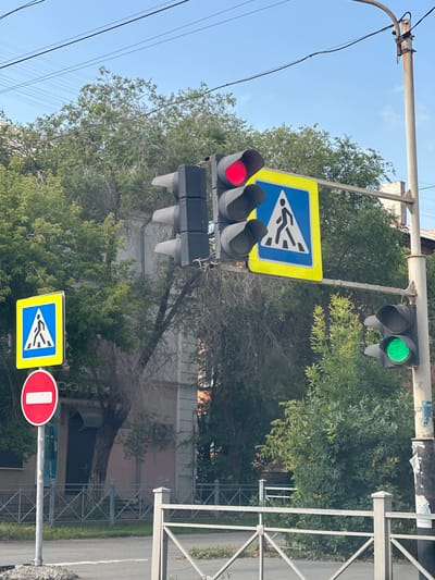 The image depicts a street scene in Orsk, Russia, featuring a traffic light with a red signal illuminated, indicating a stop. Below the traffic light, a blue square sign with a yellow border displays a white pedestrian symbol, signifying a pedestrian crossing. To the left of this setup, a red circular sign with a white horizontal bar indicates "no entry" or "do not enter." The traffic light on the right side of the image shows a green signal, suggesting that traffic can proceed in that direction. The scene is set against a backdrop of trees and buildings, with overhead power lines visible in the sky. The presence of these traffic control signs and lights suggests an intersection or a pedestrian crossing area within the city.