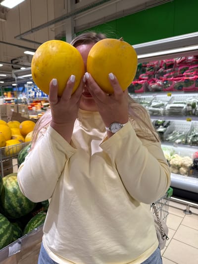 A person stands in a grocery store, holding two yellow melons in front of their face. The person is wearing a light yellow long-sleeved shirt and blue jeans. A white watch with a silver face is visible on their left wrist. In the background, shelves are stocked with produce, including watermelons, cauliflowers, and leafy greens. The store appears to be well-lit with fluorescent lighting. The scene is consistent with a retail environment, likely a supermarket or a fresh produce market. The image does not provide specific indicators to place it in Vologda, Russia, or any other particular city.