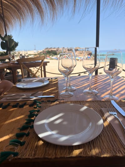 A table is set for dining at an outdoor restaurant overlooking a beach. The table is covered with a straw placemat decorated with green tassels. A white plate, a fork, and a knife are placed on the placemat. Several wine glasses are arranged on the table, and the background shows a sandy beach with people relaxing and swimming in the turquoise ocean under a clear blue sky. A thatched roof provides shade for the dining area. The location is identified as Sant Josep de sa Talaia, Spain.
