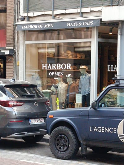The image shows a storefront on a street in Antwerp, Belgium, likely a retail establishment. The storefront has a dark awning with white lettering that reads "HARBOR OF MEN HATS & CAPS". The window display features mannequins dressed in shirts and hats, with shelves of hats visible in the background. A gray car with the license plate "2 CLA 905" is parked in front of the store. To the right of the gray car, a dark blue vehicle with the text "L'AGENCE" is also parked. The street appears to be paved with cobblestones. The building on the left has a brick facade with some windows. The environment suggests a commercial area.