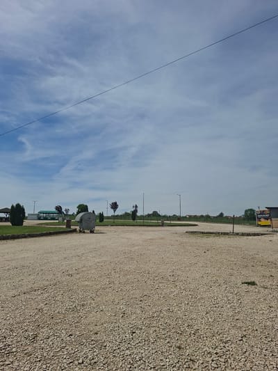 The image depicts an outdoor scene in Lukovit, Bulgaria. The foreground features a gravel-covered ground extending to the right edge. A strip of grass separates the gravel from the trees and buildings. There is a small trailer, light poles, and trees in the middle ground. The background consists of a clear sky with scattered clouds and a wire extending from the top left to the middle ground. The scene suggests a location on the outskirts of Lukovit near a parking lot or open space.