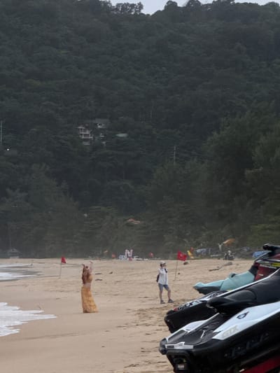 In Karon, Thailand, a beach scene unfolds with a person in a long, flowing orange skirt standing on the sand near the water's edge. The person is facing away from the camera, with their arms raised. In the mid-ground, a person wearing a white shirt, blue shorts, and a white cap stands near a red flag planted in the sand. In the foreground, several jet skis are parked on the beach, partially obscuring the sand. In the background, a dense, tree-covered hillside rises, with a few buildings visible nestled among the trees. The sky is overcast, and the overall atmosphere is muted.