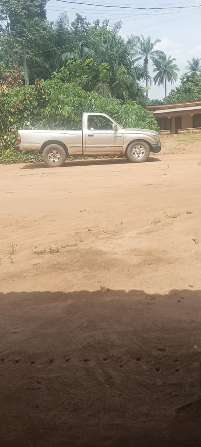 A silver Toyota Tacoma pickup truck is parked on a dirt road in Amauzu, Nigeria. Dense, tropical foliage, including palm trees, lines the background. A building with a red-tiled roof is visible behind the truck. The foreground shows the lower portion of a building, including shadows and dirt.