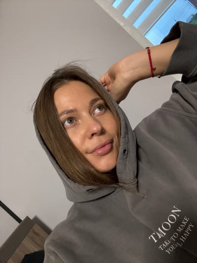 A young woman with brown hair and green eyes, wearing a grey hoodie and a red beaded bracelet, is captured in a close-up, low-angle selfie. Her right hand is raised, touching her hair, and she is looking upwards and to the right. The background is a light grey wall with a window featuring horizontal blinds, revealing a glimpse of a blue sky. A piece of furniture, possibly a bedside table with a patterned surface, is visible in the bottom left corner. The text "TMOON TAKE TO MAKE YOU'L HAPPY" is printed on the woman's hoodie. The setting appears to be indoors, potentially a bedroom, in Chernushka, Russia.