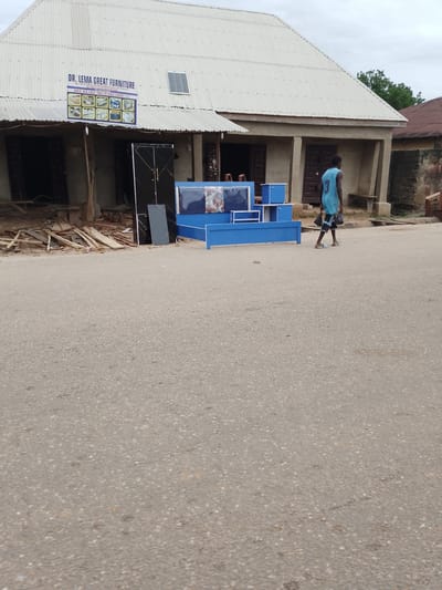 A young man walks on a paved road in front of a corrugated metal building in Serti, Nigeria. The building appears to be a furniture store, with a sign that reads "DR. LEMA GREAT FURNITURE" and shows various furniture items. In front of the building, there is blue furniture, including a bed and storage units. Wood debris is scattered on the ground. The sky is overcast. The man is wearing a light blue tank top and dark shorts, carrying two black bags in his left hand. He is walking away from the camera.
